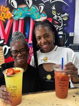 Two women smiling at the camera, holding colorful drinks at a counter with graffiti art wall in the background