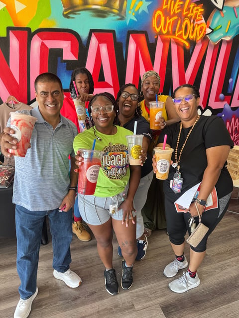 Group of six smiling people posing together in front of colorful graffiti wall art, holding drinks and wearing casual festive attire