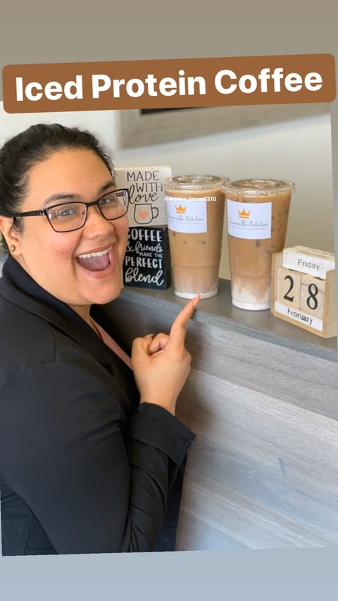 Woman smiling while pointing at two iced protein coffee drinks on counter with coffee-themed decor