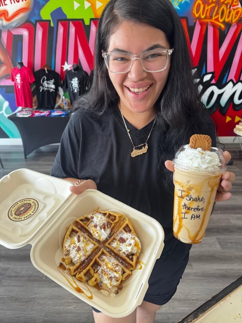 Young woman smiling at camera holding a waffle plate and iced coffee drink in a colorful cafe setting