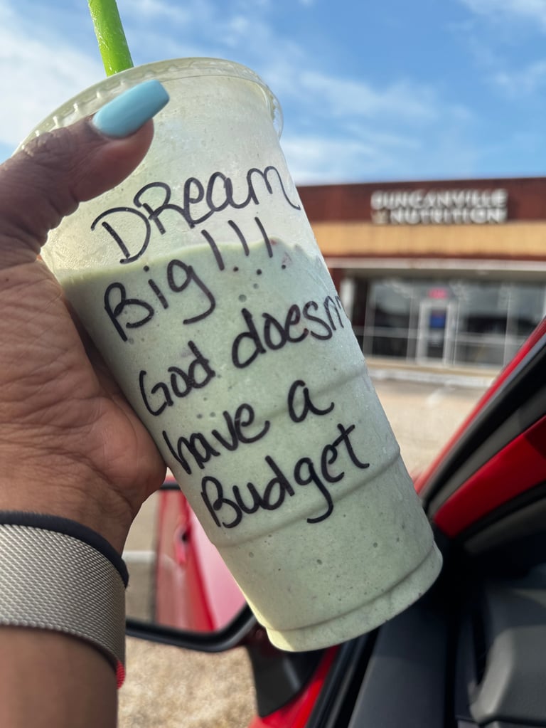 Hand holding a beverage cup with handwritten text reading Dream Big, God doesnt have a budget, outdoors